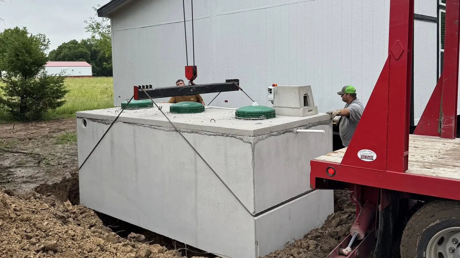 Crew guiding a precast septic tank into place from a boom truck beside a home, septic system installation