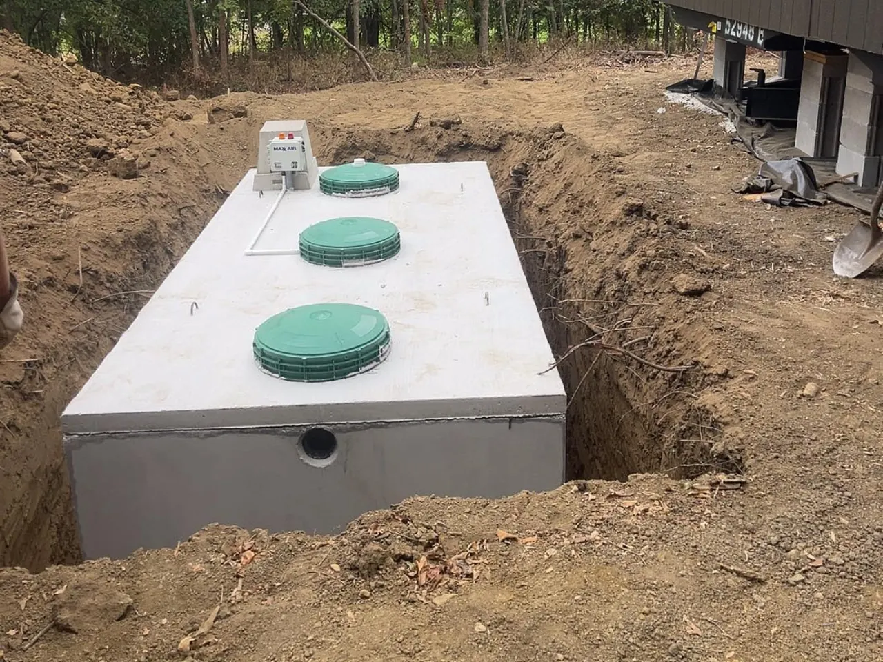 Aerobic tank with green lids and a control box set in an open hole beside a home on piers, aerobic septic systems