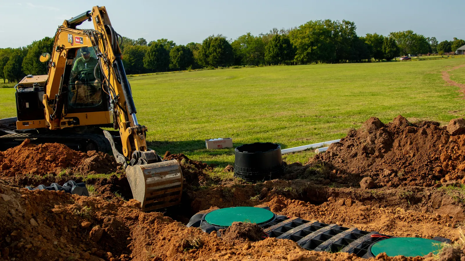 Mini excavator working beside a new plastic septic tank with green lids on an open lot, septic system installation