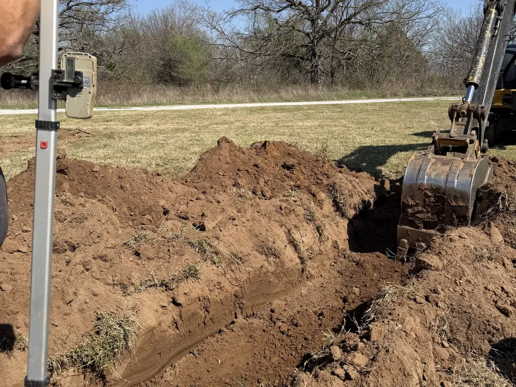 Grade rod with a laser receiver beside an excavator digging a lateral line trench, lateral line repair