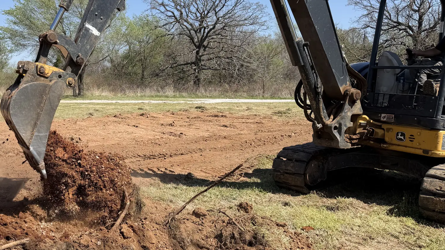 Excavator bucket dumping red clay beside a dig on an open lot, septic system installation