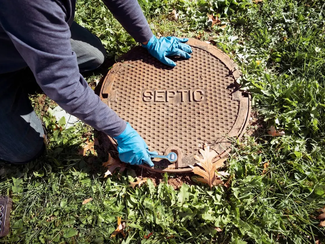 Gloved hands unbolting a cast iron septic tank lid in a grassy yard, septic inspections