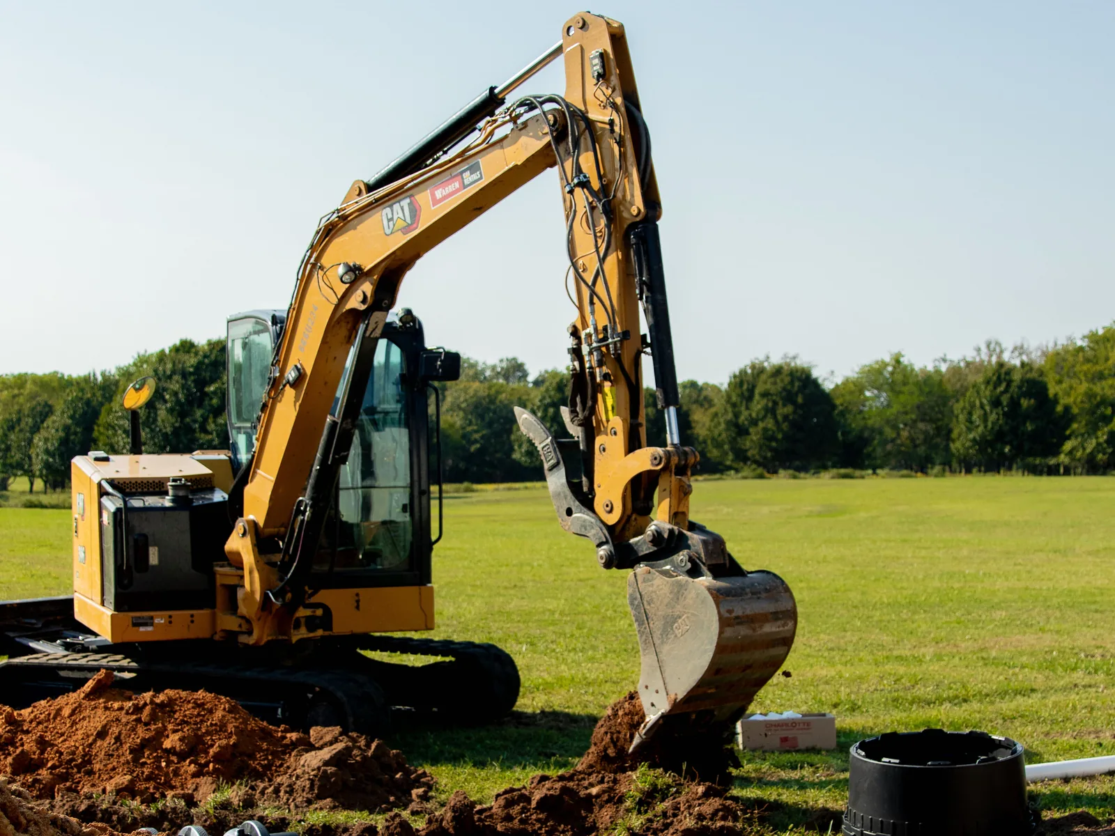 Excavator digging on an open lot beside a black tank riser, septic system installation