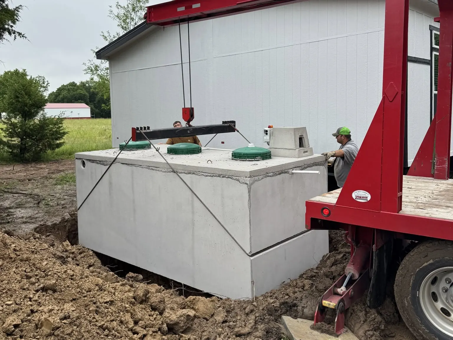Crew guiding a concrete septic tank as a boom truck lowers it into the hole beside a home, septic system installation