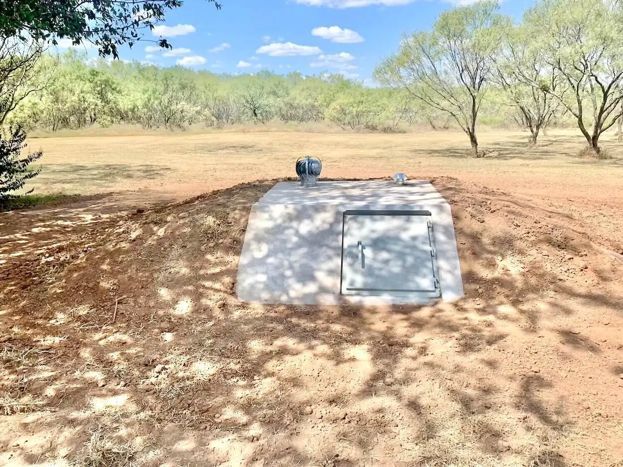 In-ground concrete storm shelter with a steel door and turbine vent set in a graded yard, storm shelters