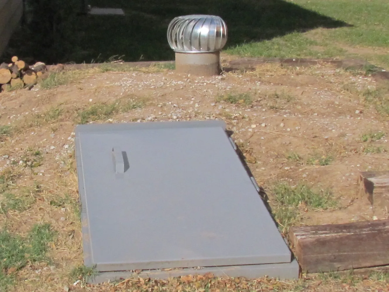 Gray steel storm shelter door and turbine vent set at ground level beside a house, storm shelter installation